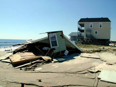 Photo showing two houses on the shoreline.  One is completely collapsed and the other appears to have only minor damage.  The standing house was elevated off the ground.